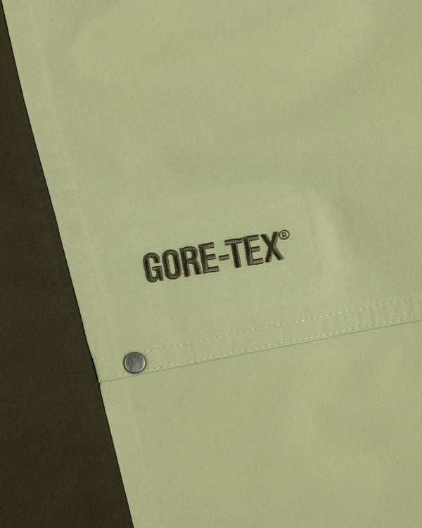 Close-up of ALD / The North Face GORE-TEX Pant: black GORE-TEX embroidery on light green fabric by a metal snap button.