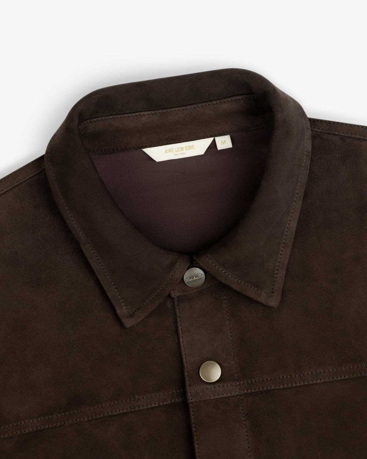 Close-up of a dark brown Suede Overshirt with a collar, buttons, and a brand label at the neckline.