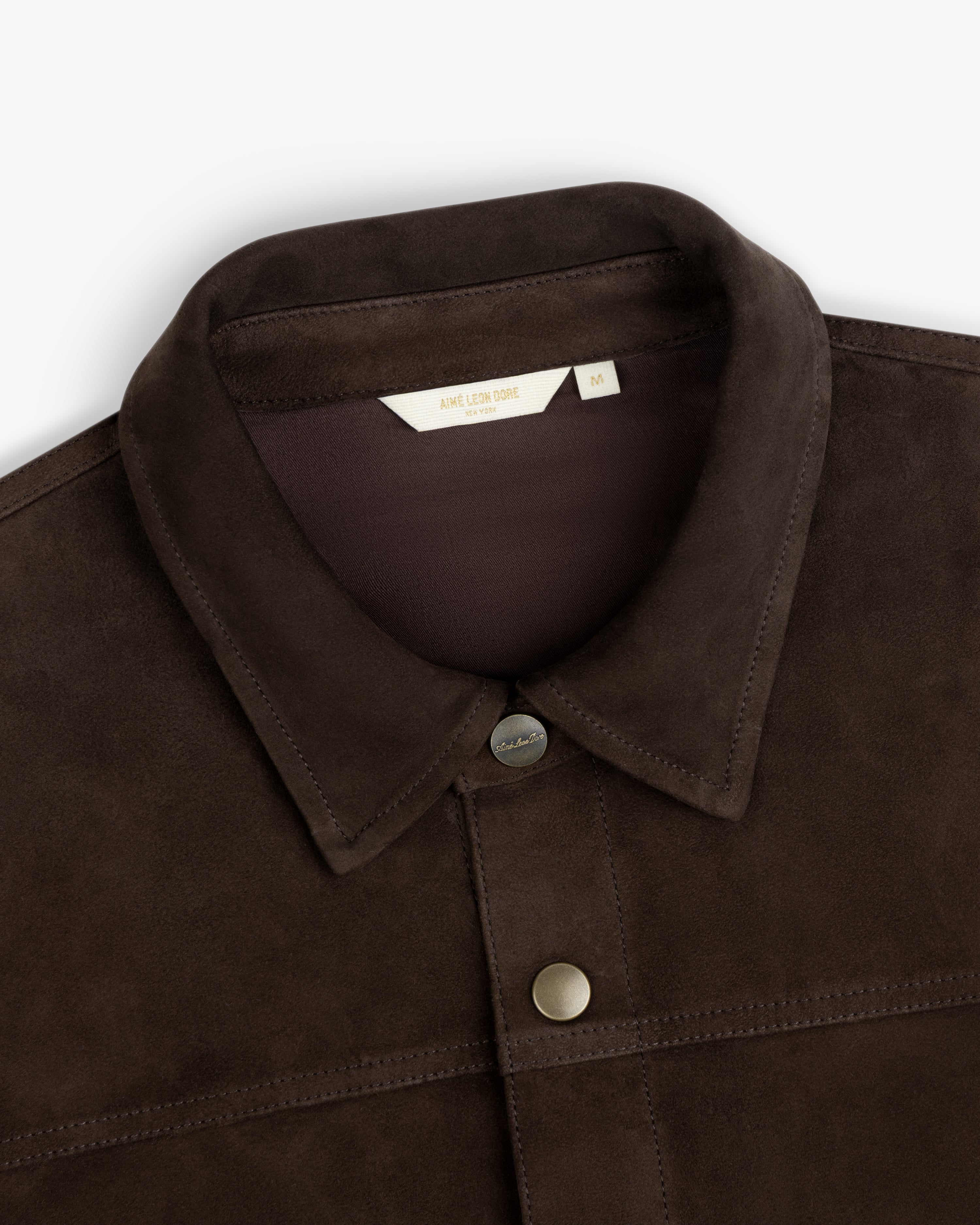 Close-up of a dark brown Suede Overshirt with a collar, buttons, and a brand label at the neckline.