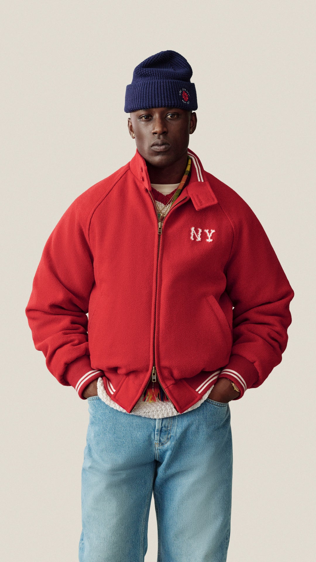 A man wearing a red varsity-style jacket with white 'NY' embroidery on the chest, paired with a navy beanie, layered necklaces, and light blue jeans, set against a solid light beige background.
