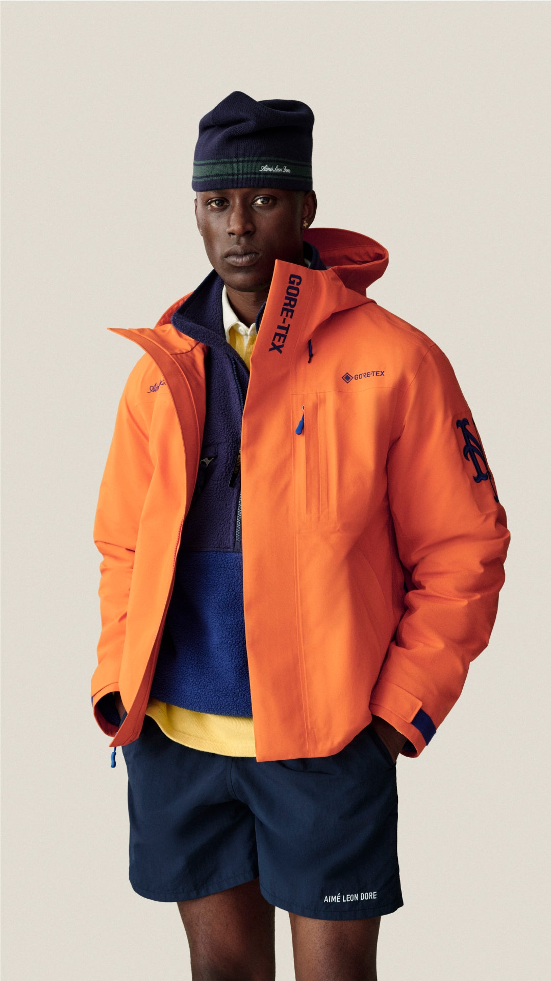 A model wearing a bright orange Gore-Tex hooded jacket over a navy sweater and yellow shirt, paired with navy shorts and a navy beanie, against a solid light beige background.