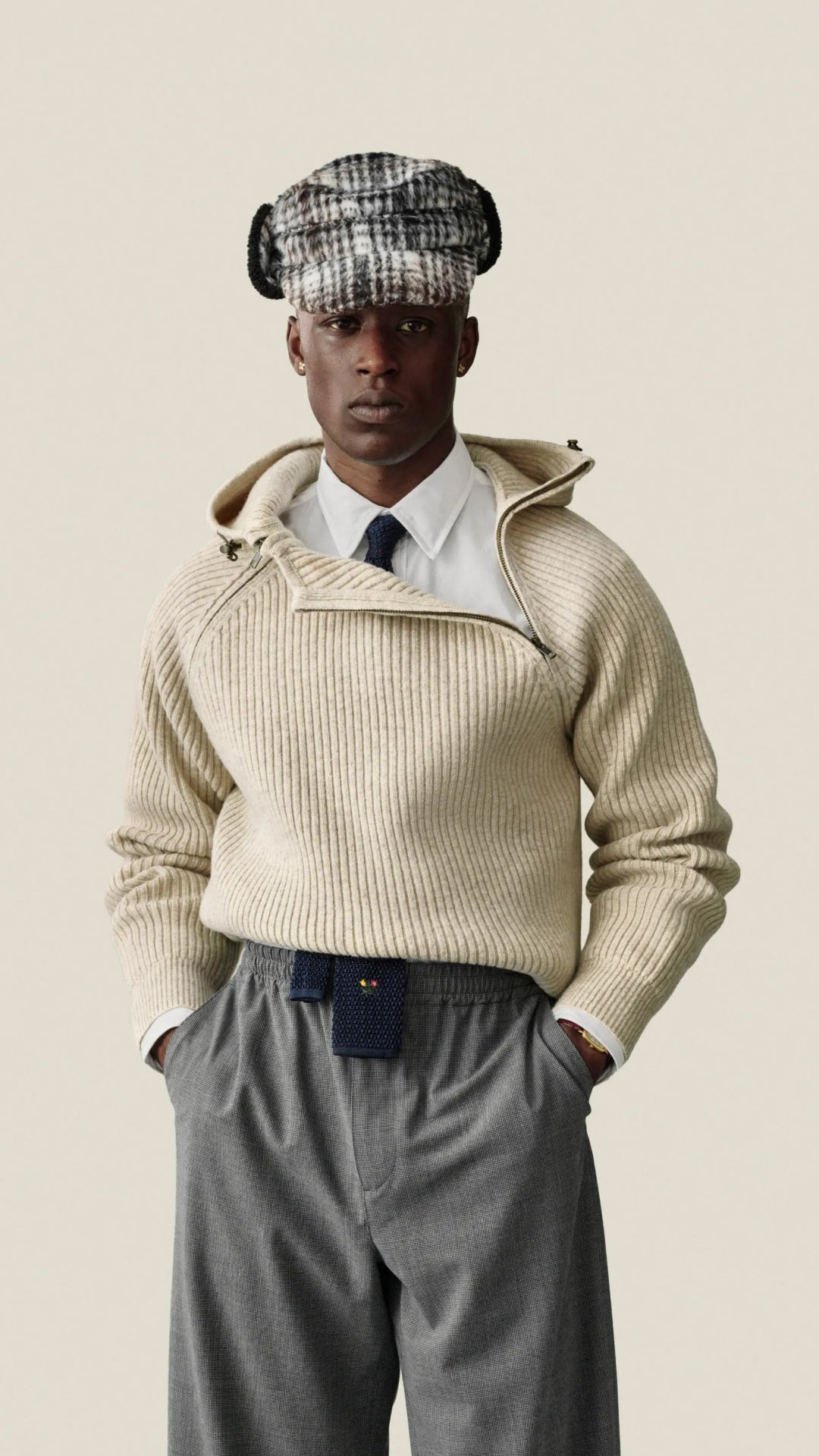 A model wearing a beige ribbed knit zip-up hoodie with a high collar, styled with a white shirt, dark tie, gray trousers, and a black-and-white checkered flat cap, set against a solid light beige background.