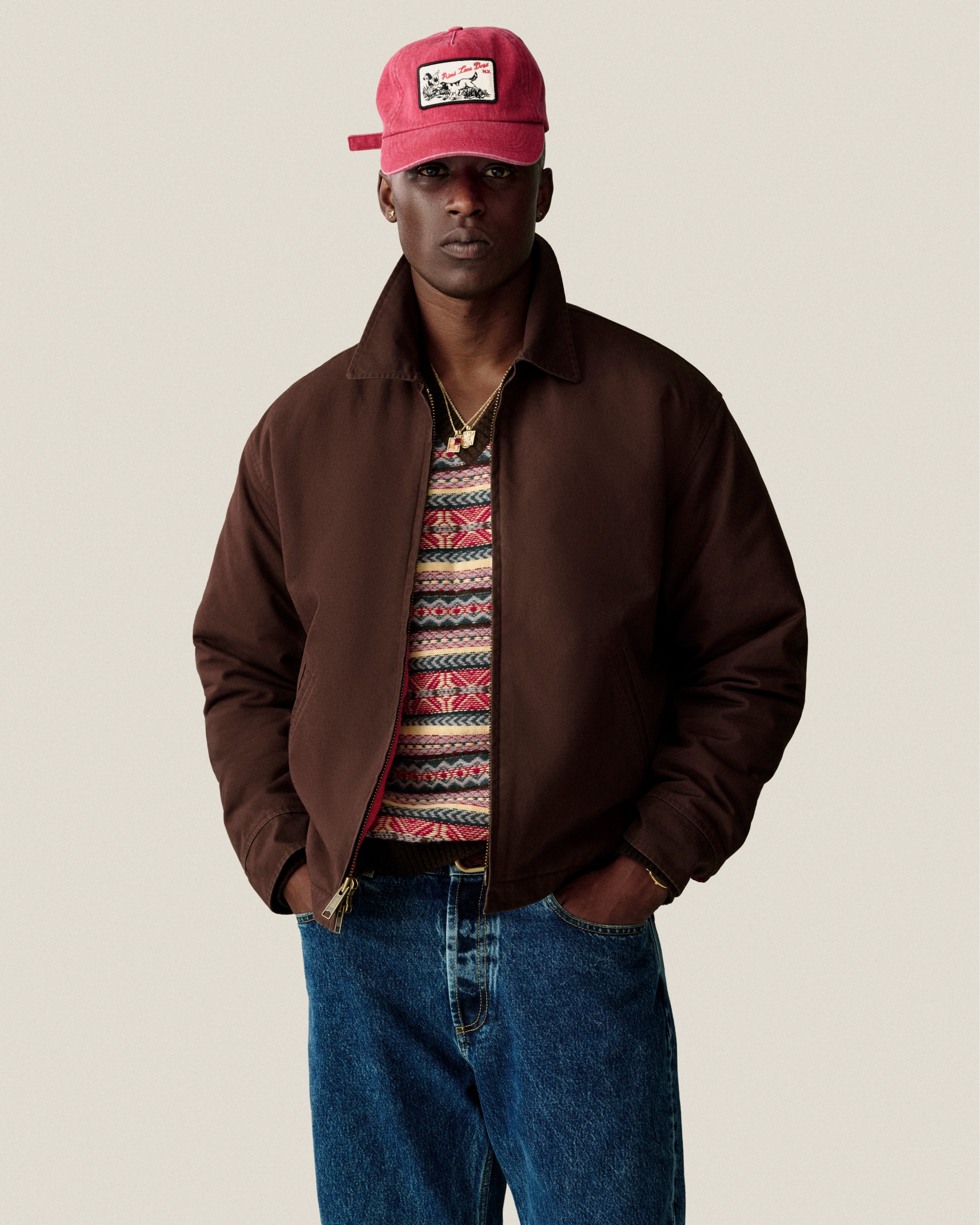 A man wears a brown jacket, Fair Isle V-Neck Sweater, blue jeans, and a red cap with patch against a plain background.