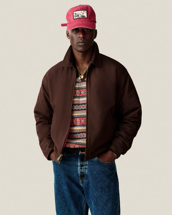 A man wears a brown jacket, Fair Isle V-Neck Sweater, blue jeans, and a red cap with patch against a plain background.