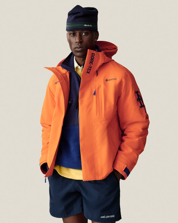 A man wears an orange GORE-TEX jacket, blue shorts, a fleece, and a Stripe Knit Beanie against a plain backdrop.