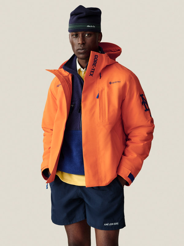 Man in an orange Mets GORE-TEX Rain Delay Jacket, blue fleece, navy shorts, and a navy beanie on a plain background.