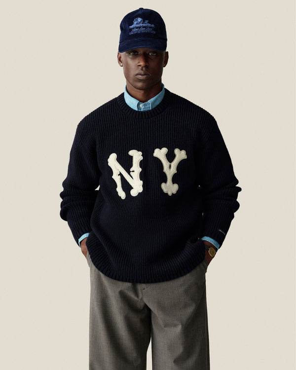 Man in navy NY sweater, blue shirt, Houndstooth Elasticated Waist Trouser, and navy cap on beige background.