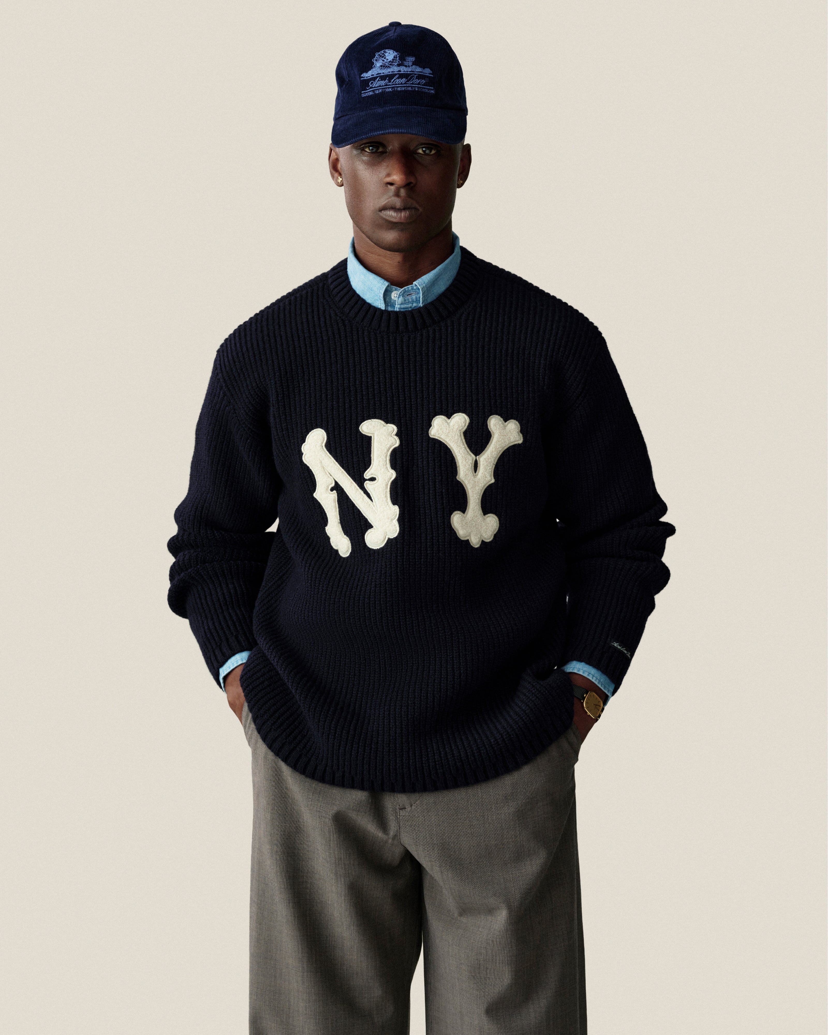 A man wears a navy NY Highlanders Ribbed Wool Crewneck Sweater, light blue shirt, gray pants, and a navy cap.