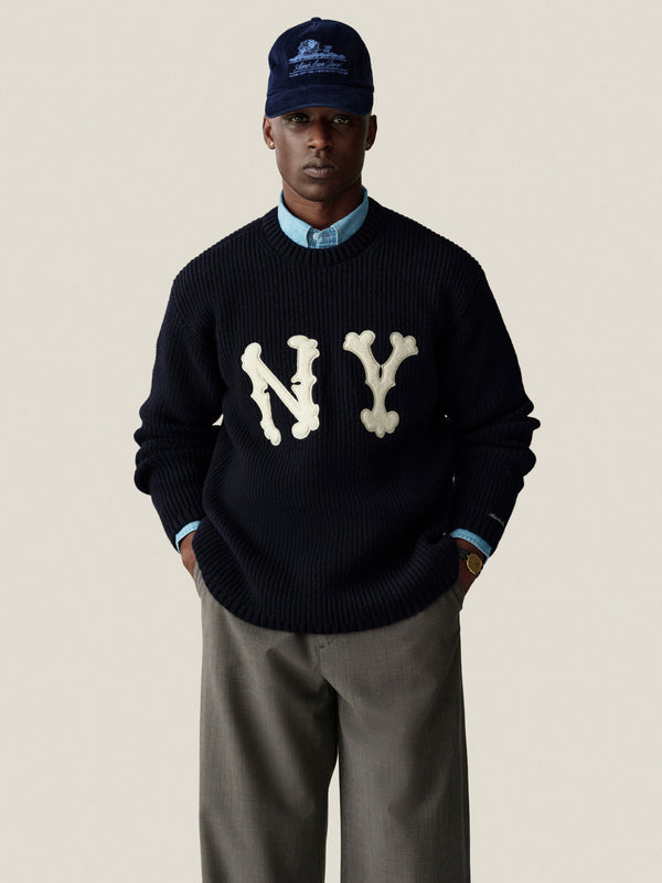 Man in NY Highlanders Ribbed Wool Crewneck Sweater, blue shirt, gray pants, and navy cap stands by a neutral backdrop.