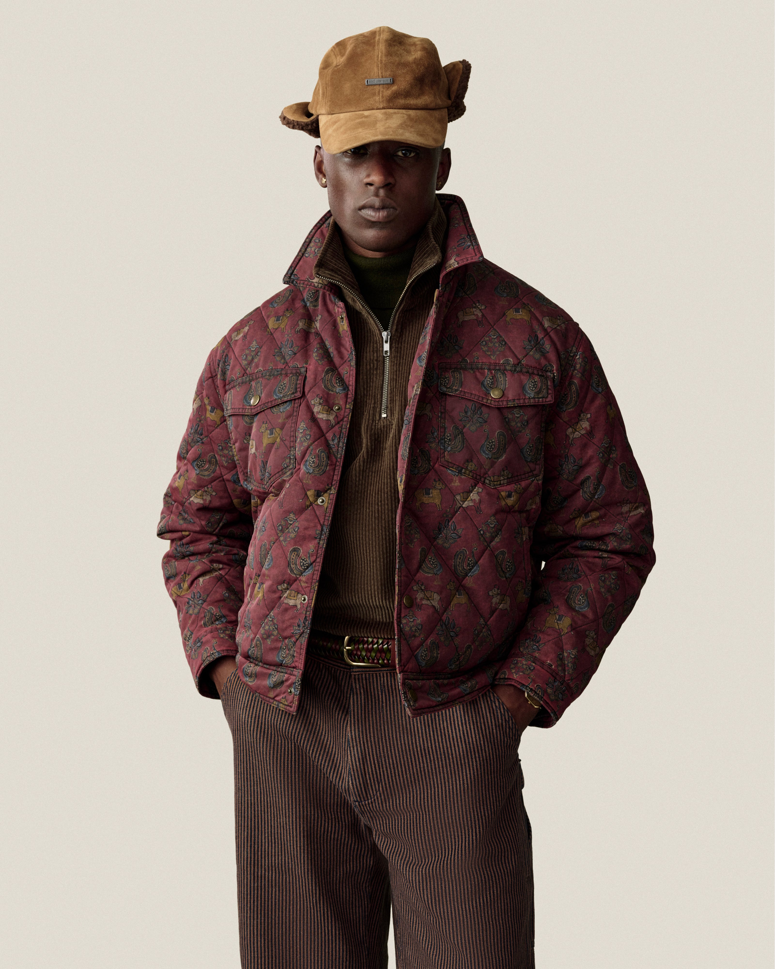 A person wears a brown Quarter Zip Corduroy Shirt under a quilted patterned jacket and a brown hat.