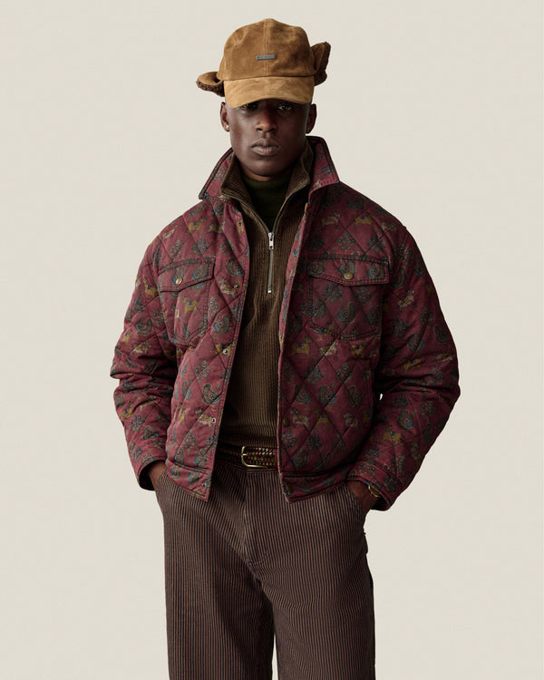 A person wears a brown Quarter Zip Corduroy Shirt under a quilted patterned jacket and a brown hat.