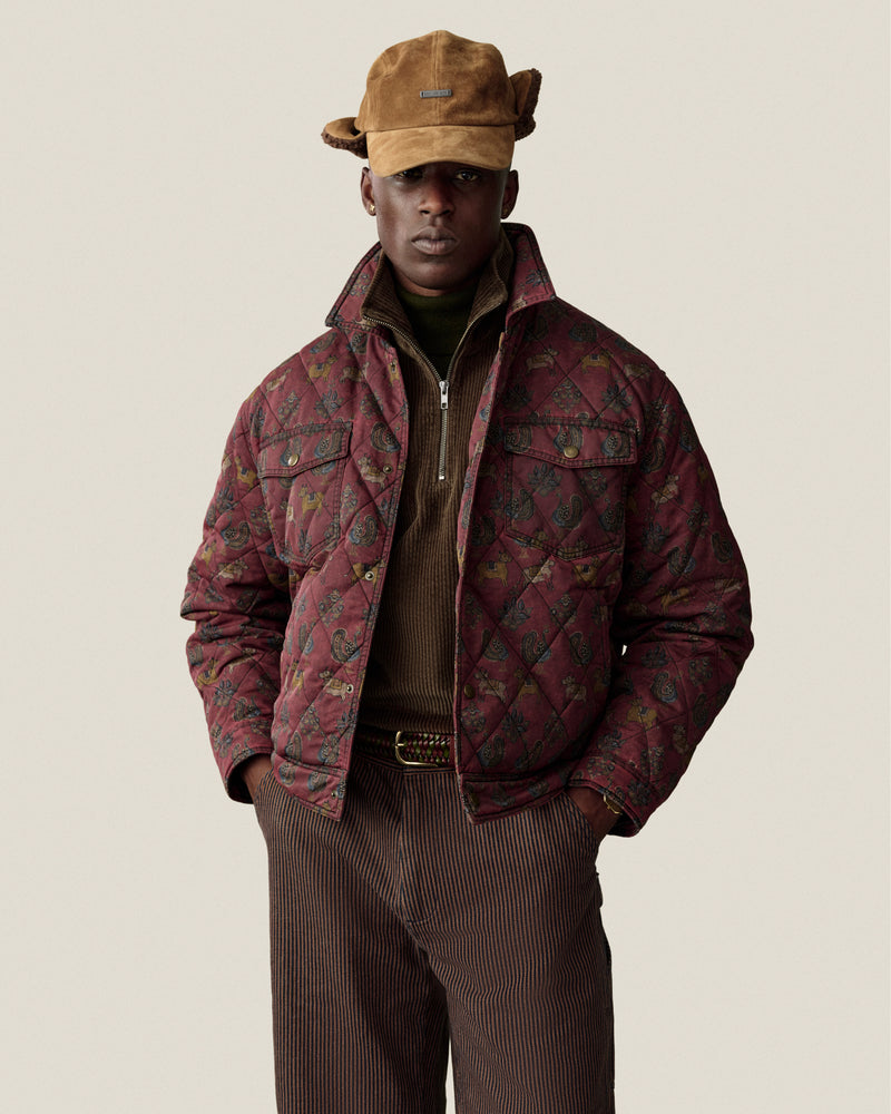 A person wears a brown Quarter Zip Corduroy Shirt under a quilted patterned jacket and a brown hat.