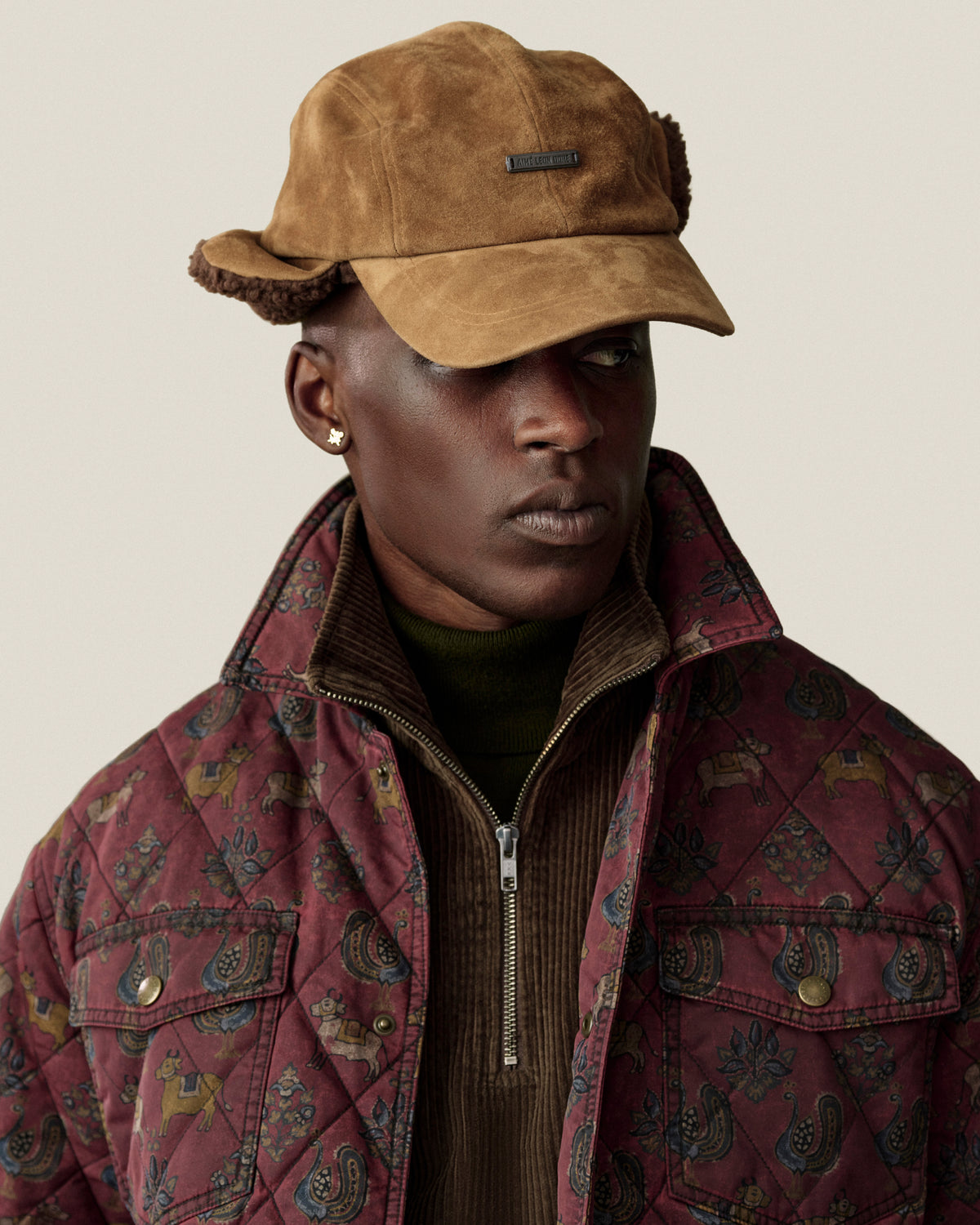A person wearing a brown suede Shearling Earflap Hat and a patterned, quilted jacket over a dark sweater.