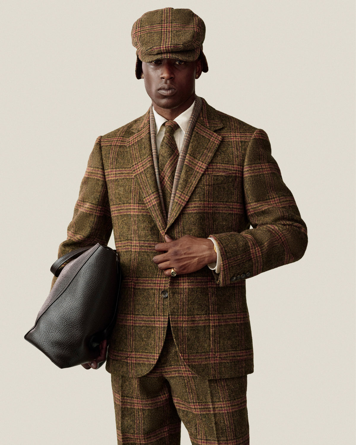 A person wearing a Single-Breasted Plaid Tweed Suit Jacket, matching cap, and tie, holding a dark leather bag.