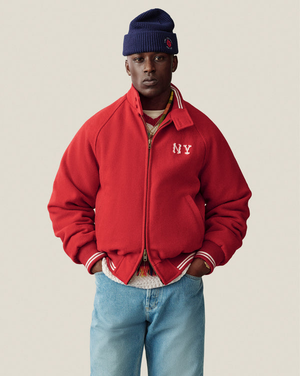 Man in NY Highlanders Wool Varsity Jacket, blue knit beanie, and jeans stands hands-in-pockets against plain background.