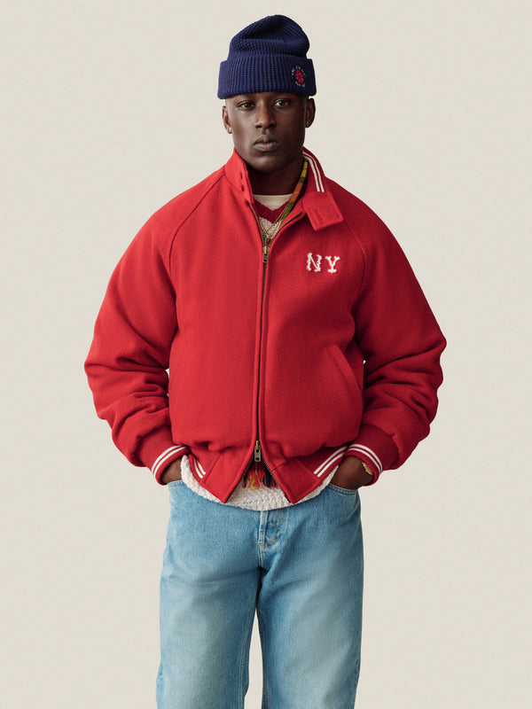 Someone wears an NY Highlanders Wool Varsity Jacket, blue knit beanie, and jeans against a plain background.