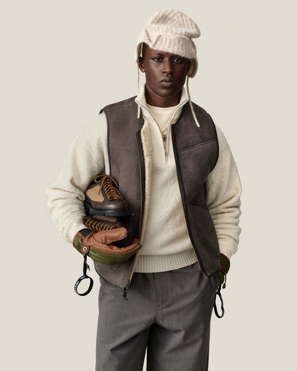 A person in a knit hat, Alpaca Wool Quarter Zip Sweater, brown vest, gray pants holds brown and black boots.
