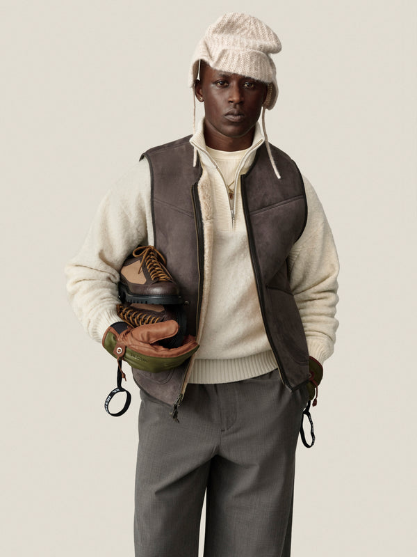 Man in an Alpaca Wool Quarter Zip Sweater, gray vest, and knit hat holds ice skates against plain background.