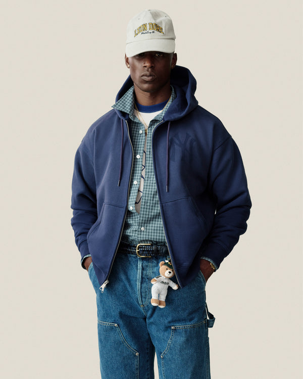 Man in blue hoodie, plaid casual shirt, jeans, beige cap, teddy bear keychain on belt, plain background.