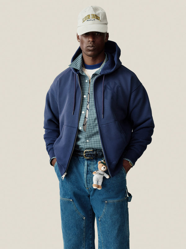Man in a Plaid Casual Shirt, blue zip hoodie, jeans, cap, necklace, and teddy bear clipped to his belt.
