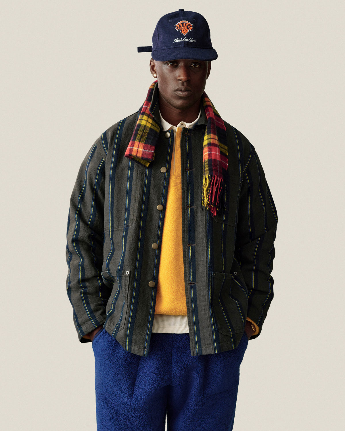 Man in Striped Denim Chore Jacket, yellow sweater, blue pants, check scarf, and navy cap against plain background.