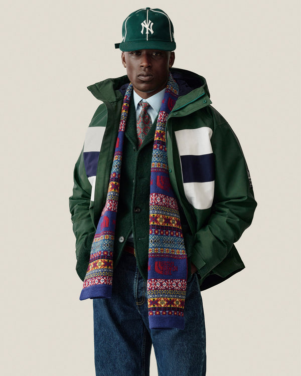 Man in GORE-TEX Striped Sailing Jacket, scarf, tie, blue jeans, and green NY cap stands against plain background.
