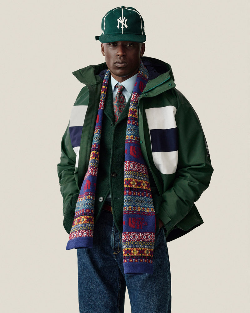 Man in GORE-TEX Striped Sailing Jacket, scarf, tie, blue jeans, and green NY cap stands against plain background.