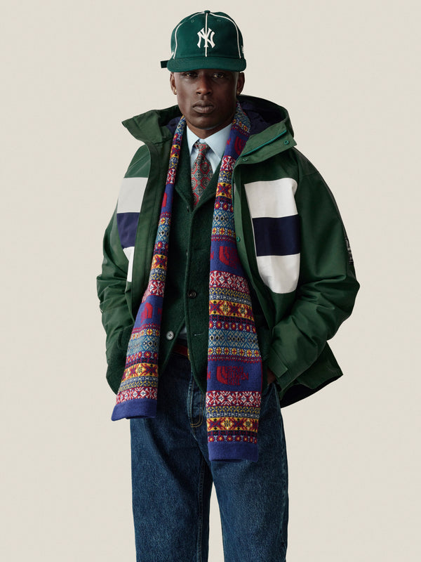 Man in green GORE-TEX Striped Sailing Jacket, colorful scarf, white shirt, tie, blue jeans on plain background.