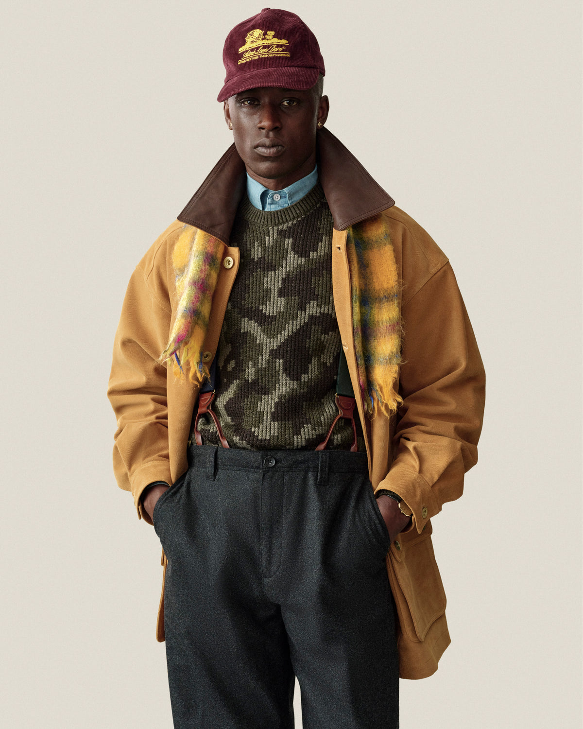 A man in a Wool Jacquard Camo Sweater, yellow plaid scarf, tan coat, and maroon baseball cap.
