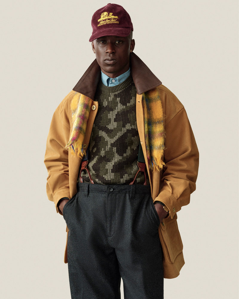 A man in a Wool Jacquard Camo Sweater, yellow plaid scarf, tan coat, and maroon baseball cap.