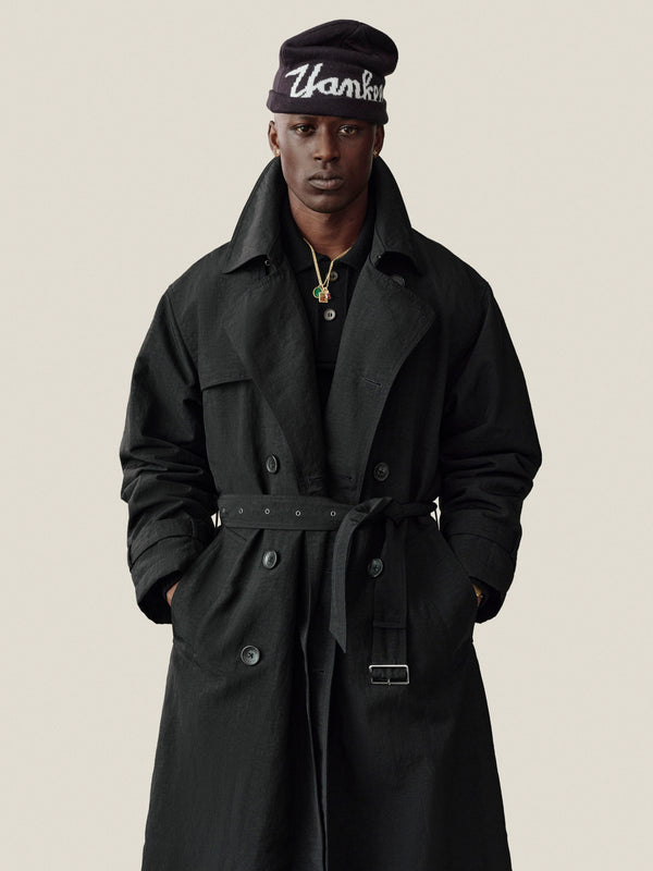 Man in a Double Breasted Nylon Trench Coat and beanie stands facing forward, hands in pockets, against a plain backdrop.
