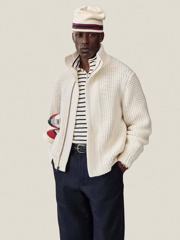 Man in a cream jacket, striped shirt, navy pants, and Stripe Knit Beanie, holding books under his arm.