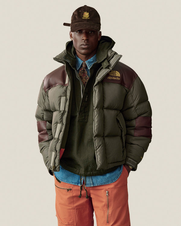 A man in a green puffer, Saint George Corduroy Hat, green shirt, blue jeans, and orange pants stands by a plain wall.