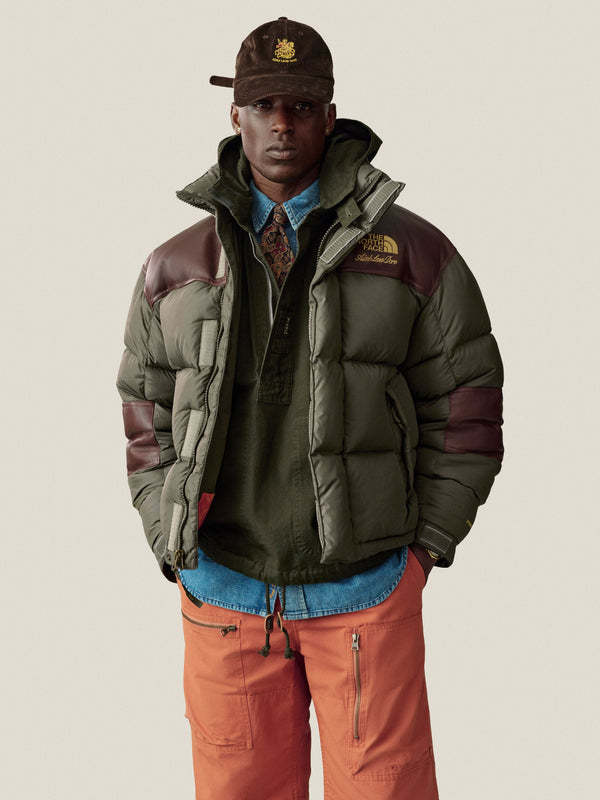 Man in a green puffer, orange pants, Saint George Corduroy Hat, layered shirts, posing against a plain background.