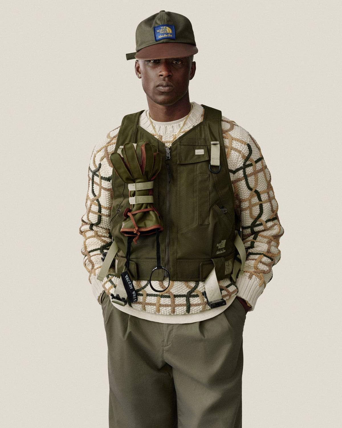 Man in a green vest, cap, and Double Pleated Officer Pant over a cream patterned sweater, against a plain backdrop.
