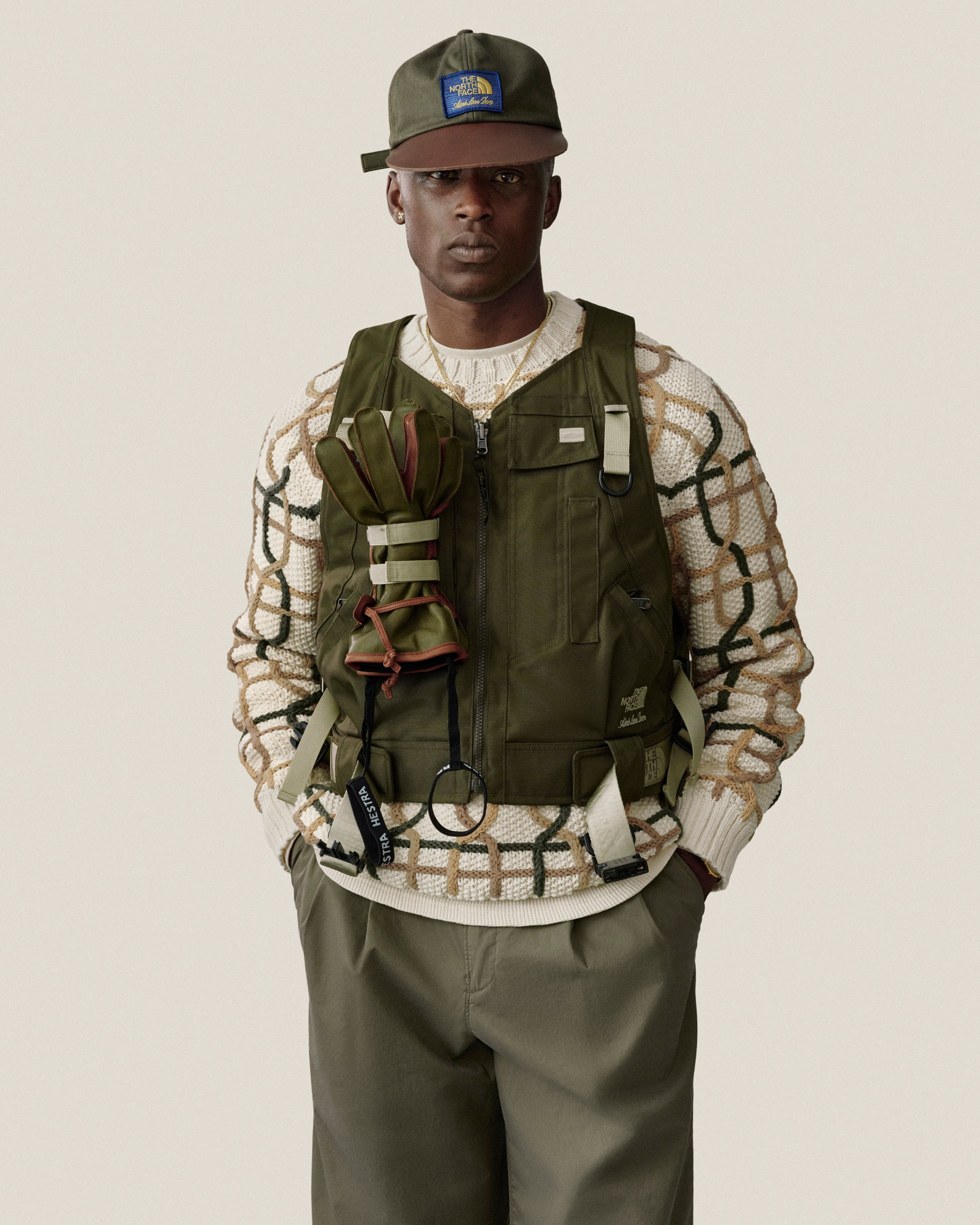 Man in a green vest, cap, and Double Pleated Officer Pant over a cream patterned sweater, against a plain backdrop.