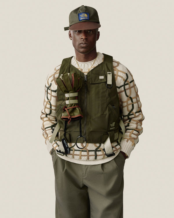 Man in a green vest, cap, and Double Pleated Officer Pant over a cream patterned sweater, against a plain backdrop.