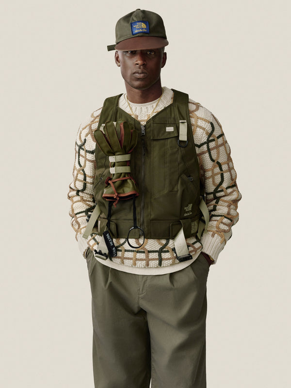 Man in Double Pleated Officer Pant, green cap and vest, over patterned sweater, stands against plain background.