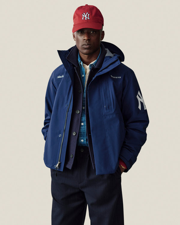 Man wears Yankees GORE-TEX Rain Delay Jacket, red cap, blue shirt, and dark pants against a plain background.