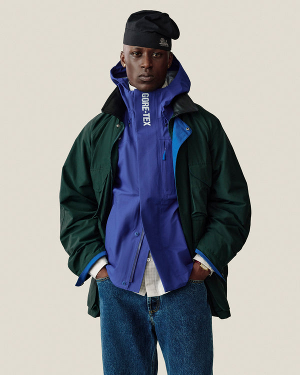 A person in a Dodgers GORE-TEX Rain Delay Jacket, black cap, green coat, and jeans stands with hands in pockets.