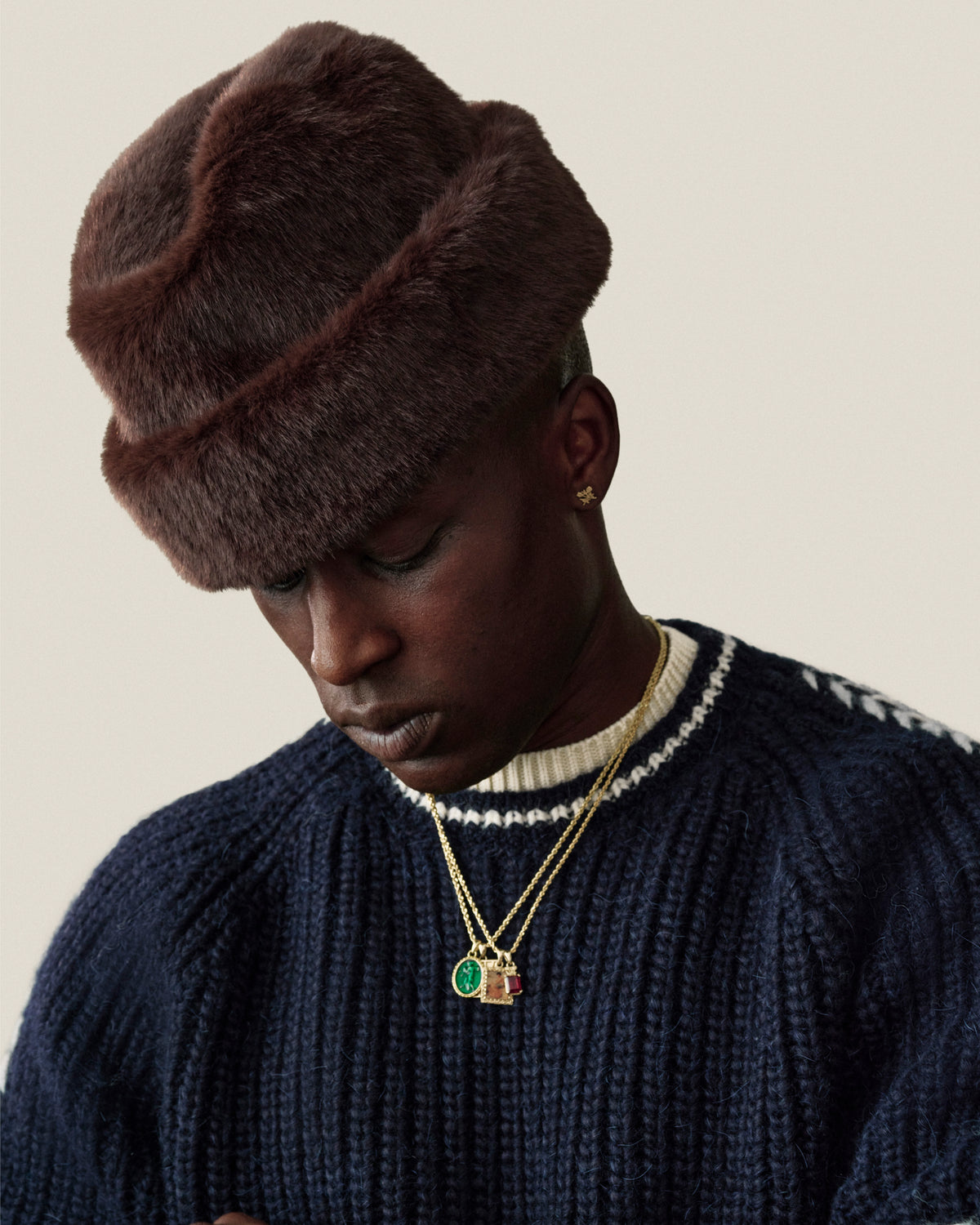A person wearing a dark brown Fur Envoy Hat and a navy blue ribbed sweater with layered gold necklaces.