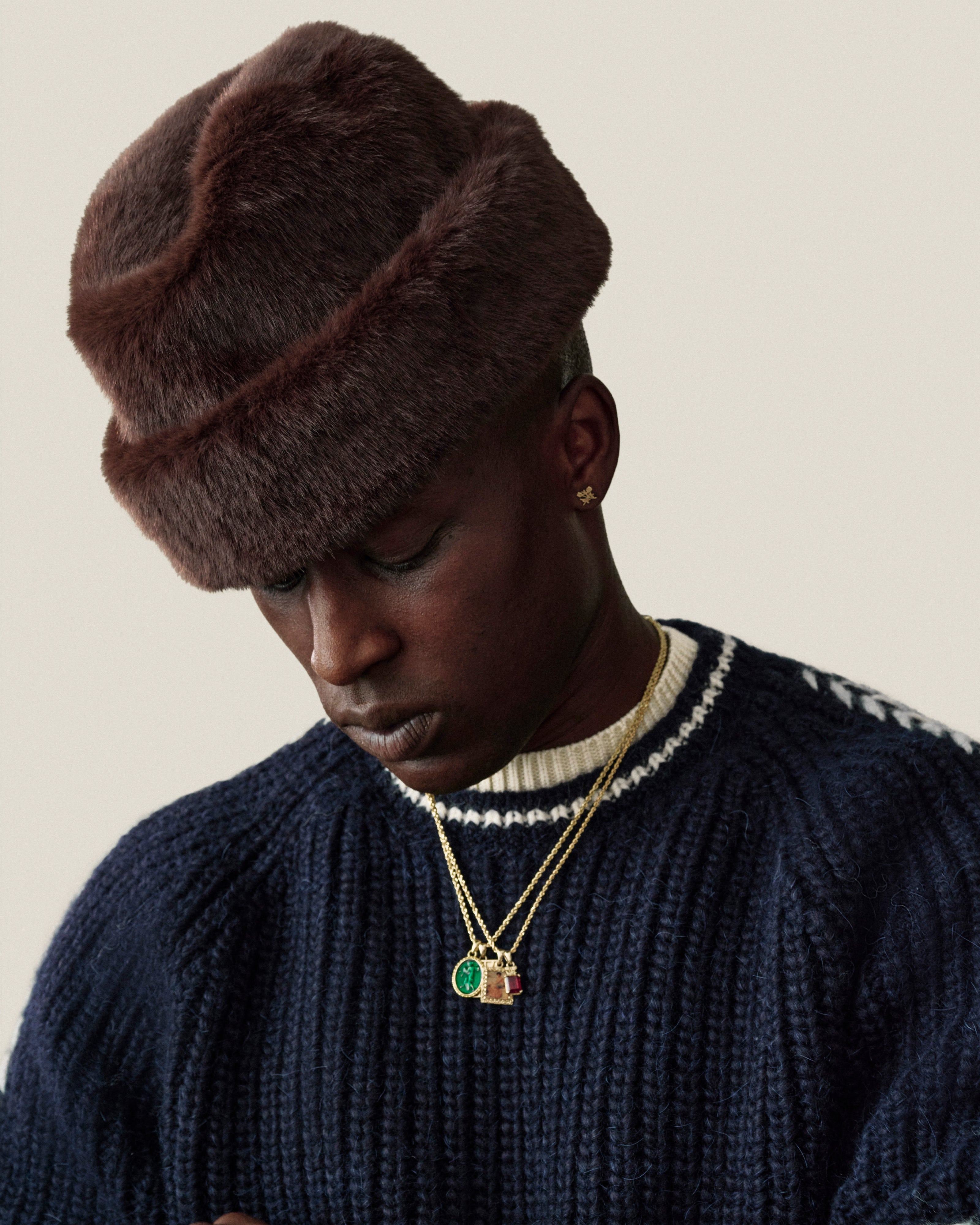A person wearing a dark brown Fur Envoy Hat and a navy blue ribbed sweater with layered gold necklaces.