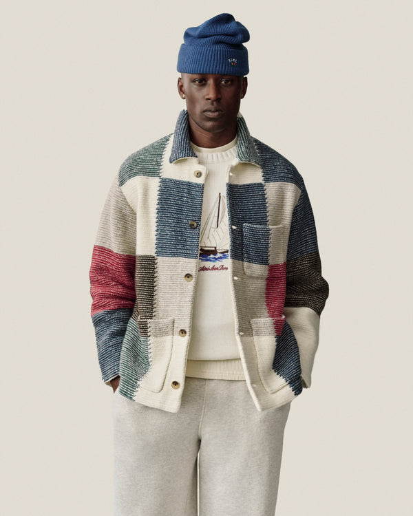 Man in patchwork jacket, Regatta Embroidered Crewneck Sweater, gray pants, and blue beanie stands against plain background.