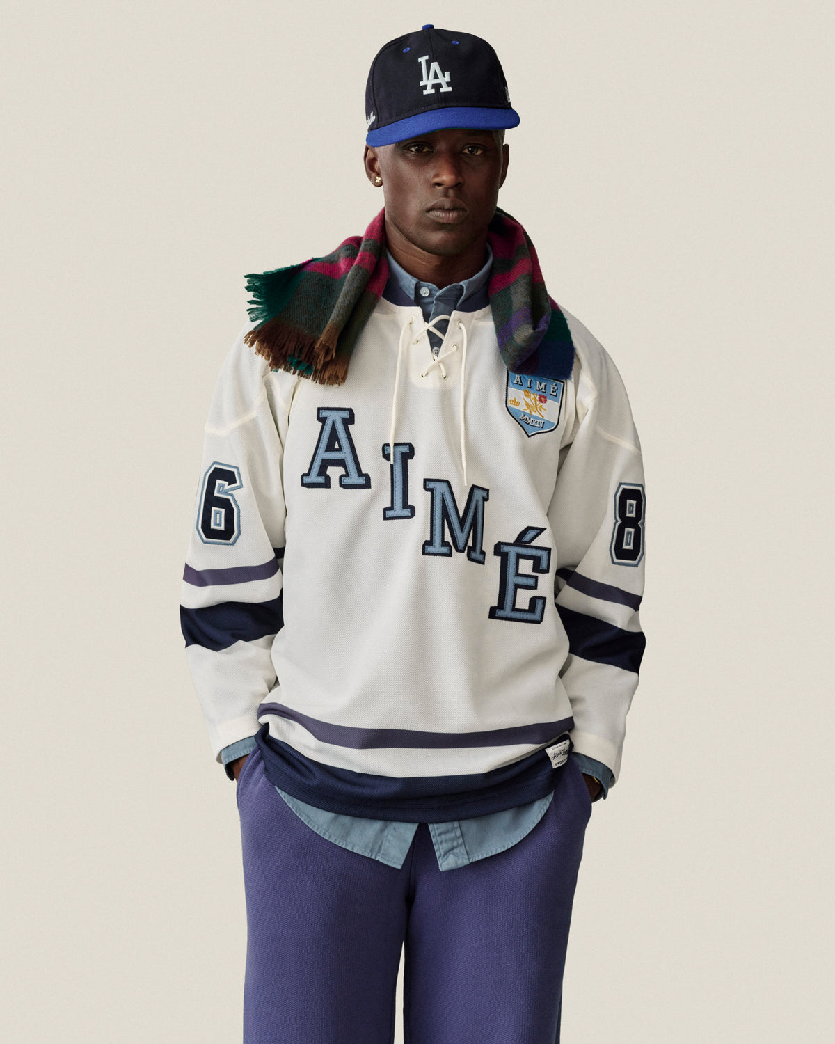 Man in Stone Washed Marina Sweatpants, LA cap, plaid scarf, AIMÉ 86 white hockey jersey, hands in pockets.