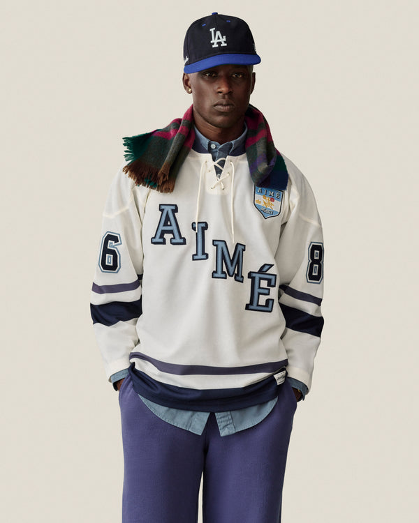 Man in Stone Washed Marina Sweatpants, LA cap, plaid scarf, AIMÉ 86 white hockey jersey, hands in pockets.