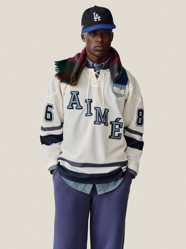 A man wears a cream AIMÉ hockey jersey, Stone Washed Marina Sweatpants, plaid scarf, and a blue LA cap.