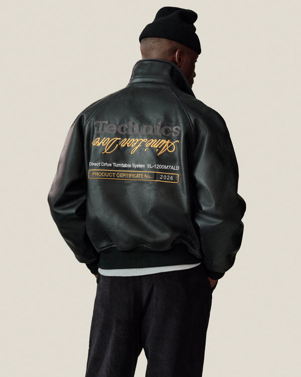 A person in the ALD / Technics Varsity Leather Jacket and a black beanie stands facing away, text embroidered on back.
