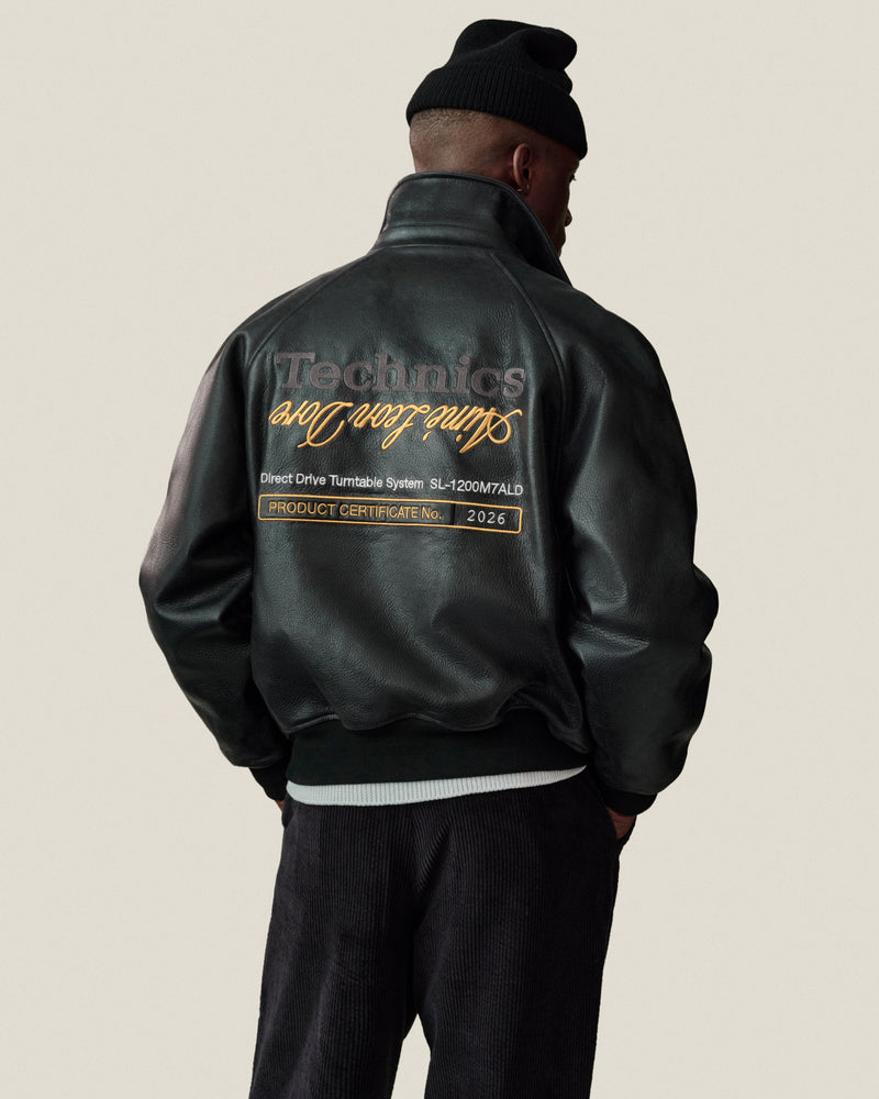 A person in the ALD / Technics Varsity Leather Jacket and a black beanie stands facing away, text embroidered on back.
