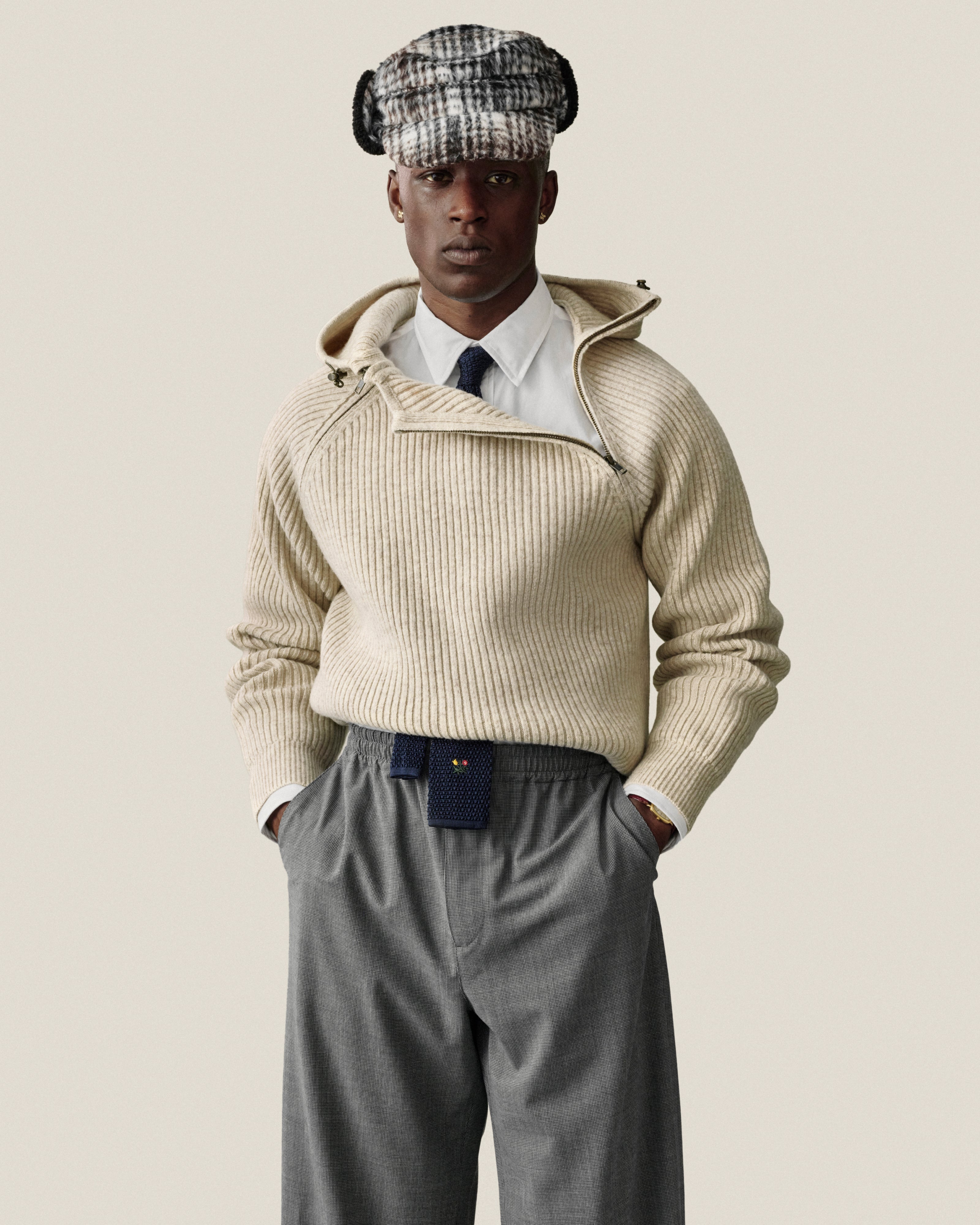 Person in a cream Wool Ribbed Knit Hoodie with an asymmetrical zipper, white shirt, tie, grey trousers, and a plaid cap.