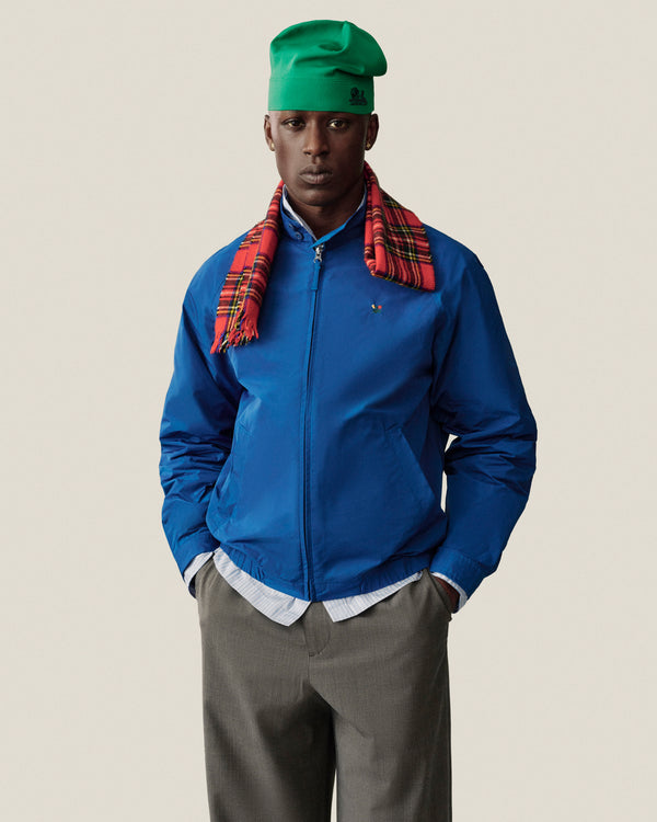 Man in Crest Nylon Windbreaker, gray pants, green hat, red plaid scarf stands with hands in pockets against plain background.